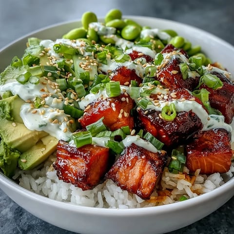 A close-up of a Salmon Rice Bowl showing baked salmon cubes, fluffy rice, edamame, and avocado with spicy mayo drizzle.